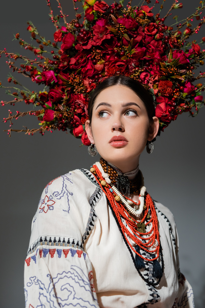 portré barna ukrán nő hagyományos ruházat és virágos vörös koszorú néz félre elszigetelt szürke - Fotó, kép