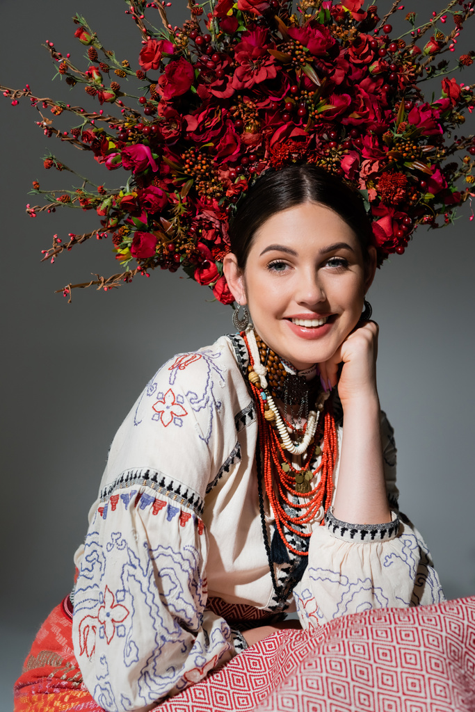 portrait of happy ukrainian woman in traditional clothing and floral red wreath isolated on grey - Foto, imagen