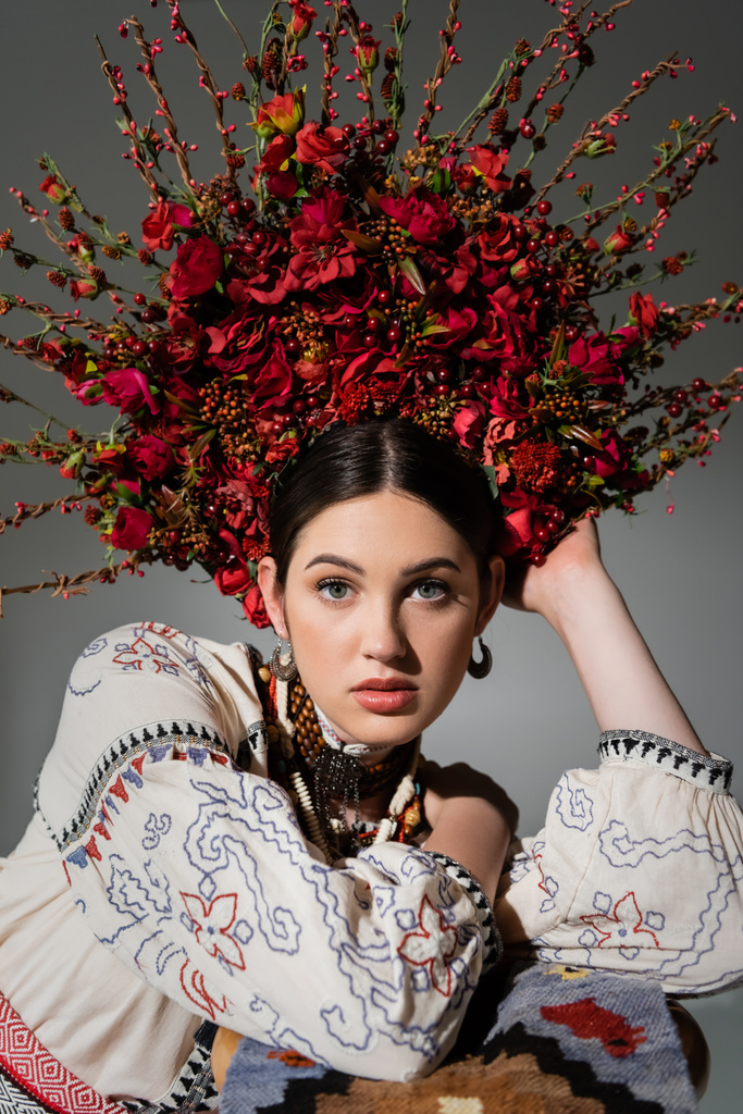 portrait of ukrainian woman in traditional clothes and floral wreath with red berries looking at camera isolated on grey - Photo, image