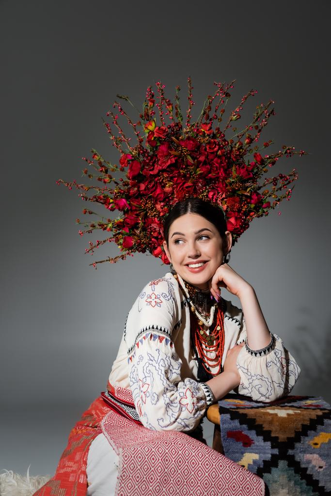 ευτυχισμένη Ουκρανή γυναίκα με παραδοσιακά ρούχα και λουλουδάτο κόκκινο στεφάνι σε γκρι - Φωτογραφία, εικόνα