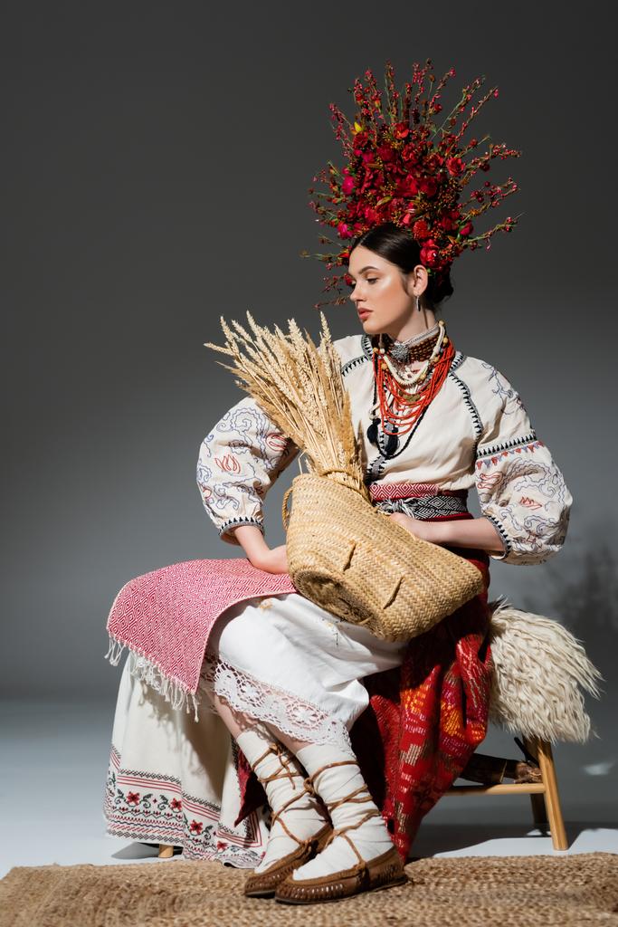 pleine longueur de jeune femme ukrainienne en couronne rouge avec des fleurs tenant sac avec du blé sur gris foncé - Photo, image
