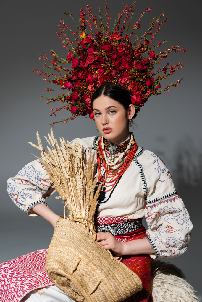 pretty ukrainian woman in traditional clothes and red wreath with berries holding bag with wheat on grey - Foto, Imagem