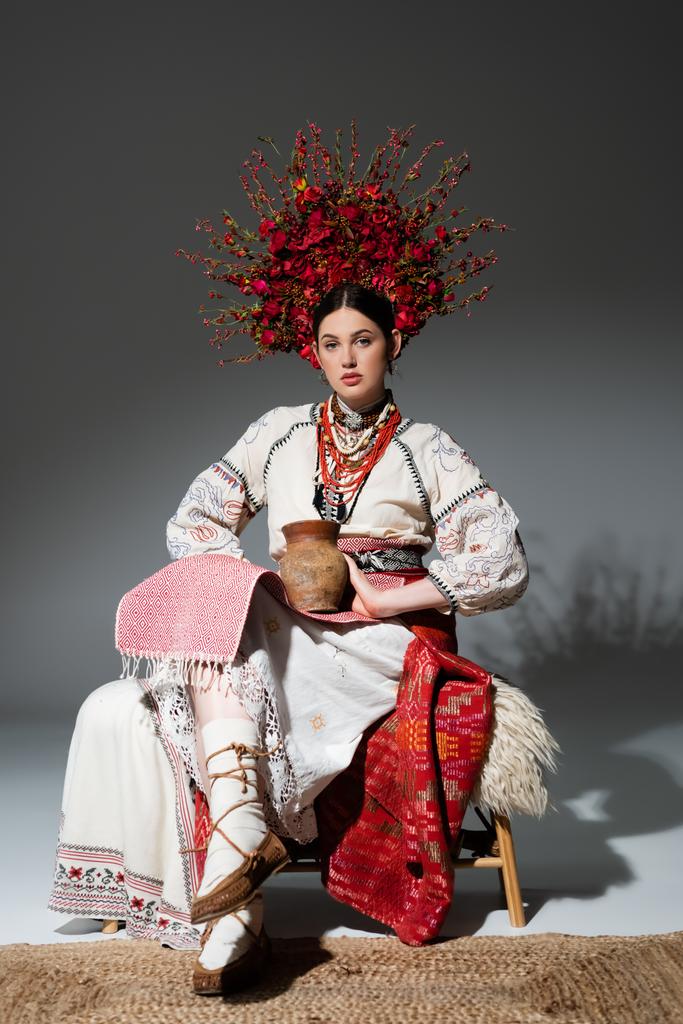 full length of young ukrainian woman in red wreath with flowers and berries holding clay pot on dark grey - Foto, imagen