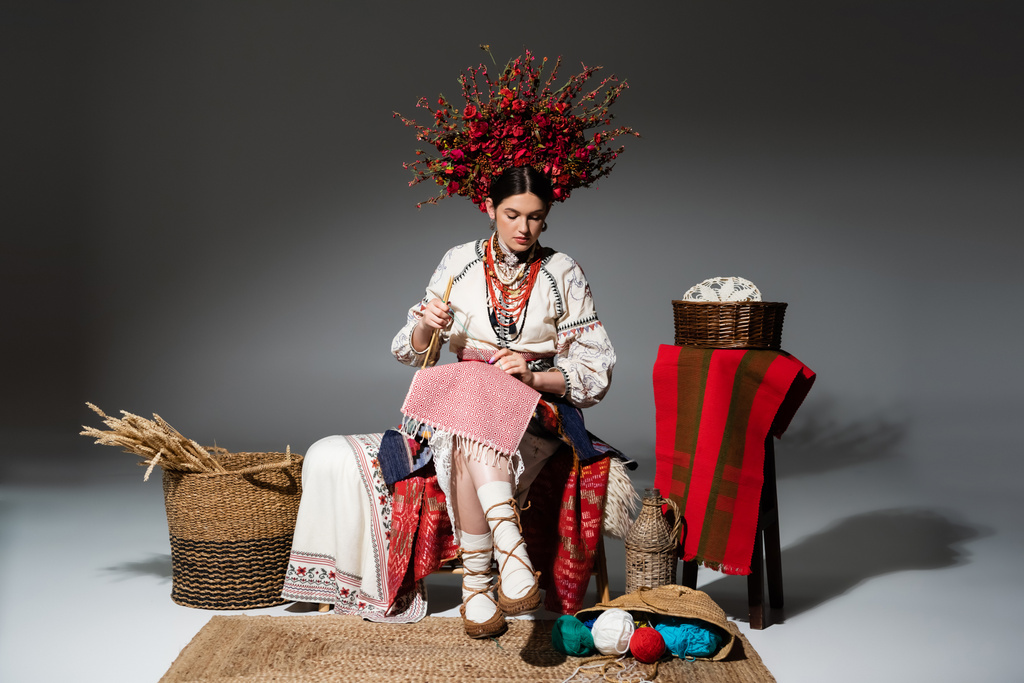 full length of ukrainian woman in traditional clothes and red floral wreath holding knitting needles on dark grey - Foto, Bild