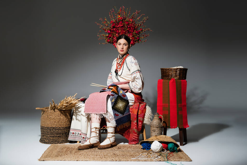 full length of young ukrainian woman in traditional clothes and red floral wreath holding knitting needles on dark grey - Valokuva, kuva