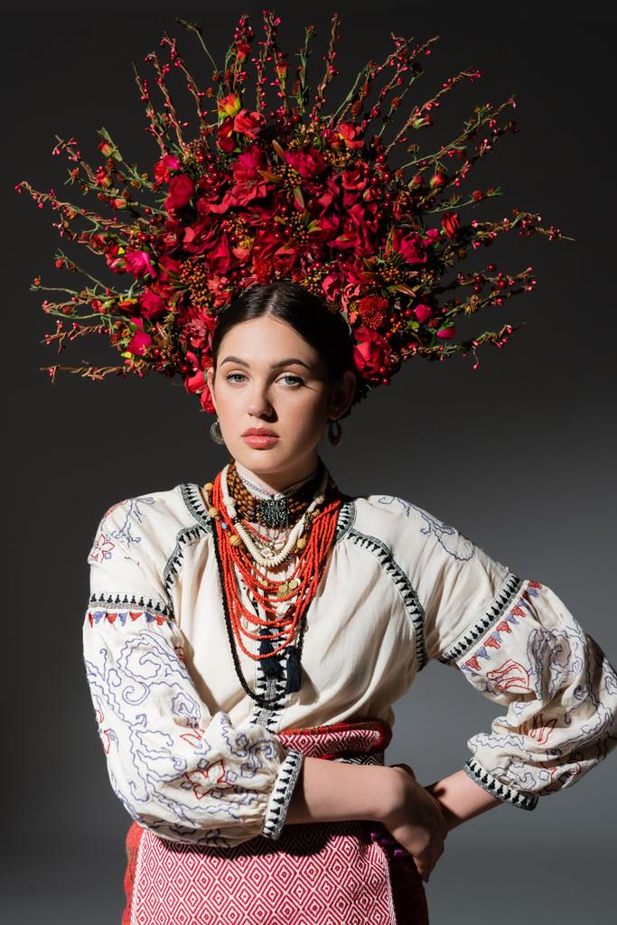 brunette and young ukrainan woman in floral wreath with red berries posing with hand on hip on dark grey - 写真・画像