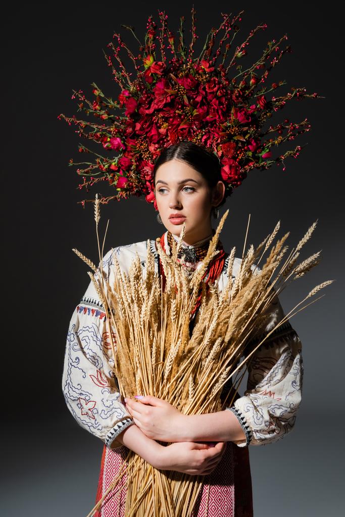 украинская женщина в цветочном венке с красными ягодами держит пшеничные шипы на темно-сером - Фото, изображение