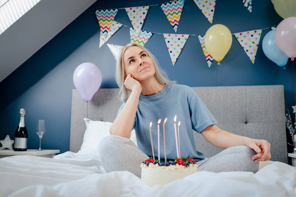 Thoughtful smiling woman in birthday party cap with festive cake with candles on the bed in decorated bedroom. Make a wish process. Morning surprise. Happy birthday concept. Selective focus. - Photo, Image