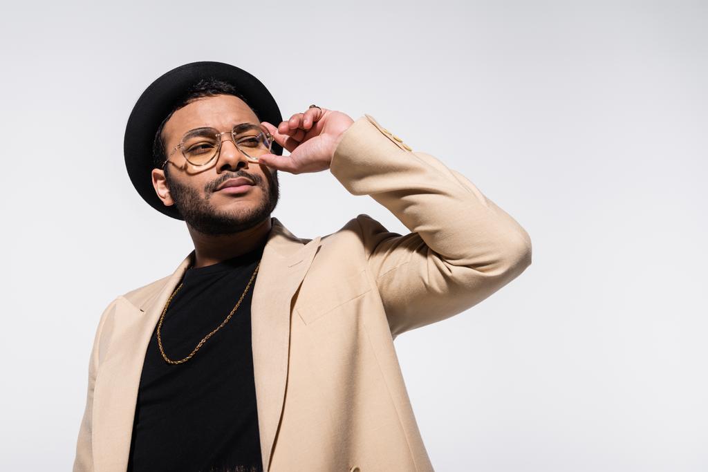 стильный индийский хип-хоп исполнитель в шляпе fedora регулируя очки изолированы на сером - Фото, изображение