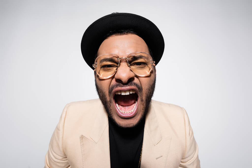 emotional indian hip hop performer in fedora hat and eyeglasses screaming isolated on grey - Zdjęcie, obraz