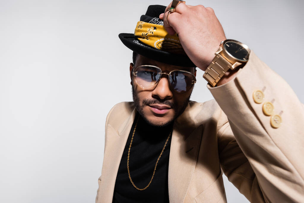 eastern hip hop performer with golden wristwatch holding fedora hat and looking at camera isolated on grey - Photo, Image