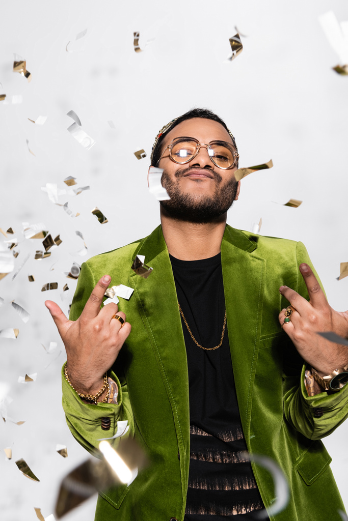confident indian hip hop performer in green velvet blazer and crown near falling confetti on grey - 写真・画像