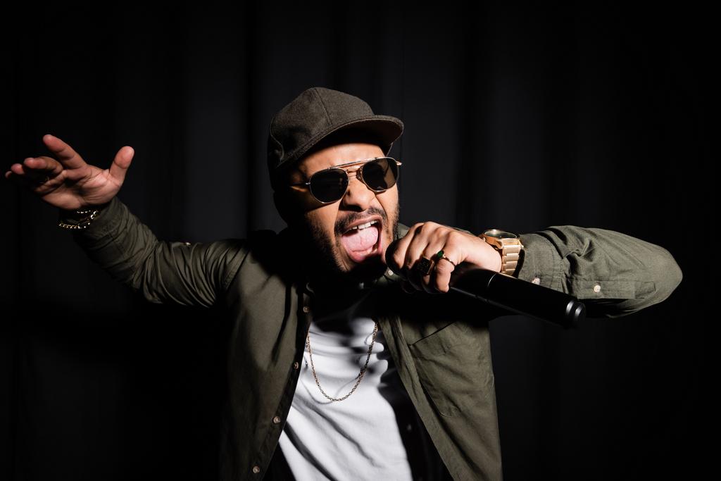 oostelijke hiphop zanger in zonnebril zingen in microfoon op zwart - Foto, afbeelding