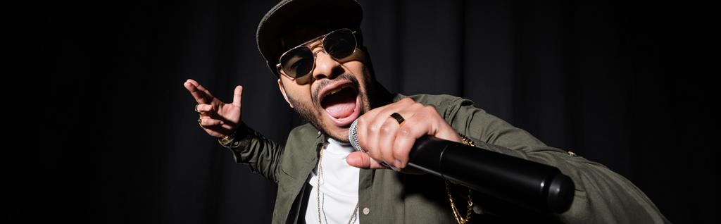 indian hip hop performer in sunglasses and cap singing in microphone on black, banner - Photo, Image