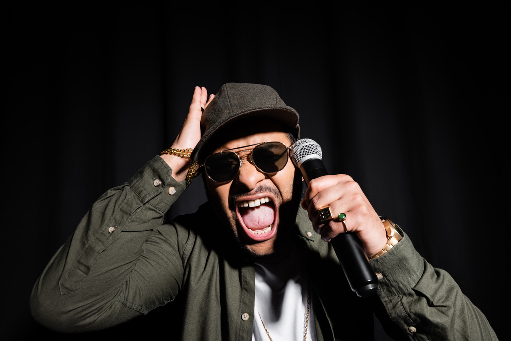 emotional middle east hip hop performer in sunglasses and cap screaming while holding microphone isolated on black - Foto, Bild