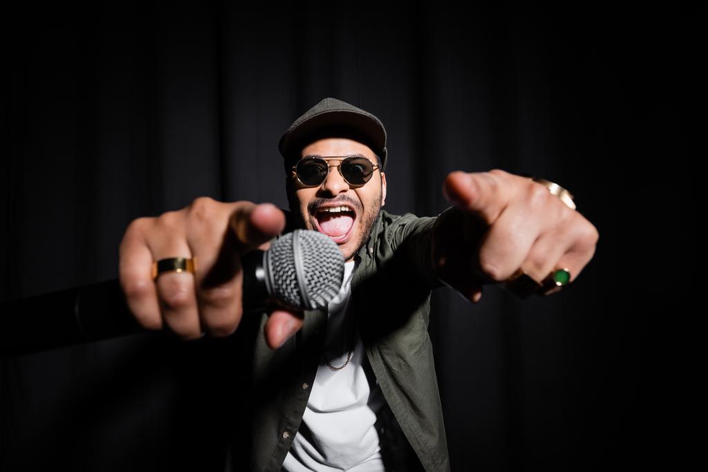 excited middle east hip hop performer in sunglasses and cap singing in microphone and gesturing on black - Zdjęcie, obraz