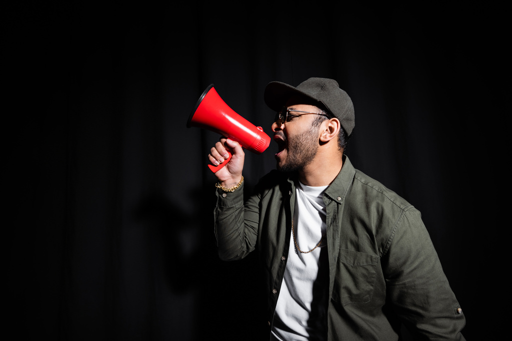 Seitenansicht eines emotionalen Hip-Hop-Performers aus dem Nahen Osten mit Sonnenbrille und Schirmmütze, der singt und Lautsprecher auf schwarz hält - Foto, Bild
