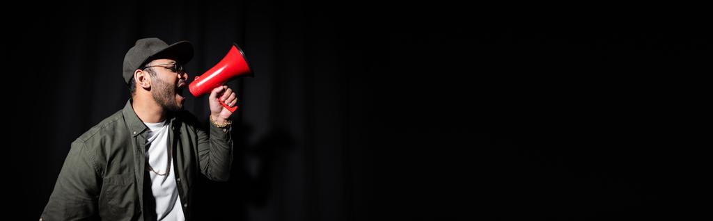 вид сбоку эмоционального исполнителя хип-хопа Среднего Востока в солнцезащитных очках и кепке, поющего и держащего громкоговоритель на черном баннере - Фото, изображение
