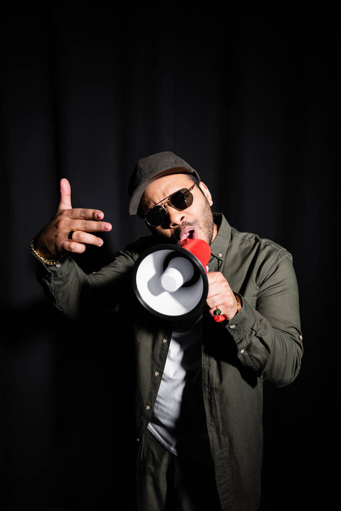 emotional middle east hip hop performer in sunglasses and cap talking in megaphone on black - Photo, Image