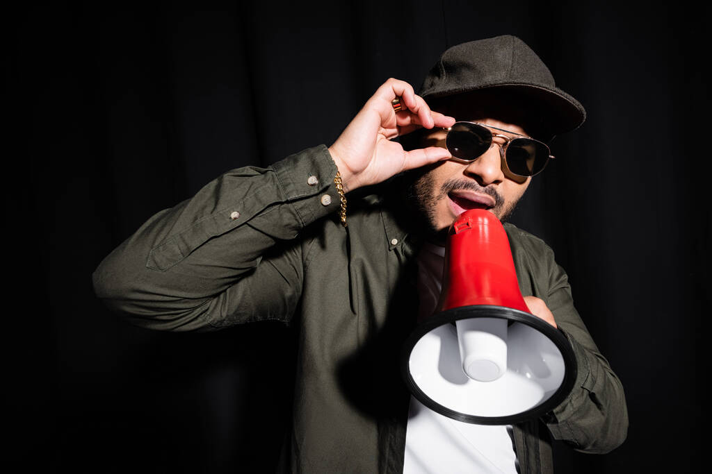 Emotionaler Hip-Hop-Performer aus dem Nahen Osten setzt Sonnenbrille auf und spricht im Lautsprecher auf schwarz - Foto, Bild