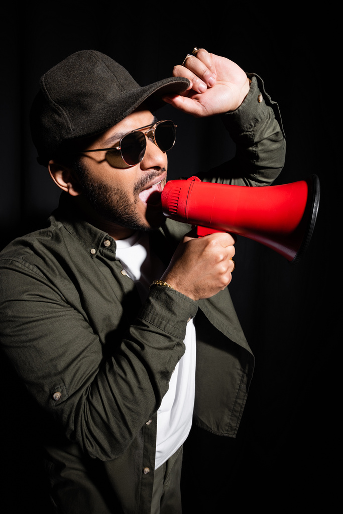 indian hip hop singer in sunglasses and cap screaming in megaphone on black - Zdjęcie, obraz