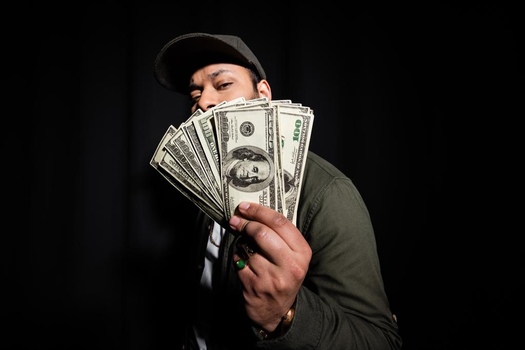 бородатый индийский хип-хоп исполнитель в кепке покрывает лицо долларовыми банкнотами на черном  - Фото, изображение