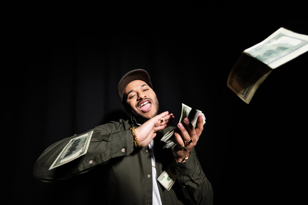 excited and rich indian hip hop performer in cap throwing dollar banknotes on black  - Fotó, kép