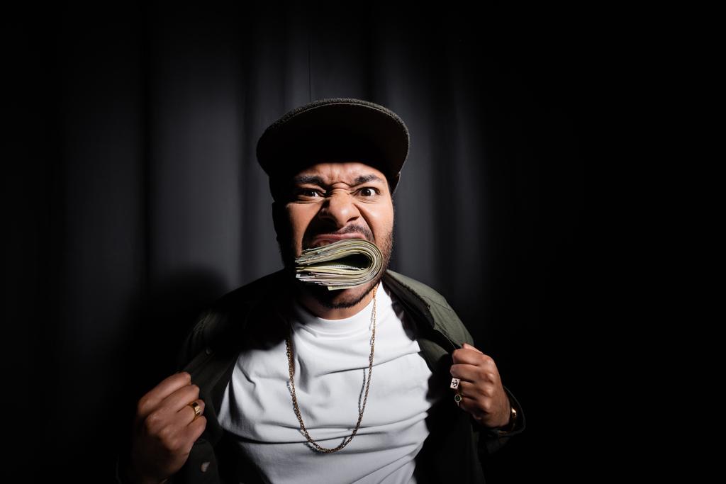 индийский хип-хоп исполнитель в кепке кусая связку долларовых банкнот на черном  - Фото, изображение