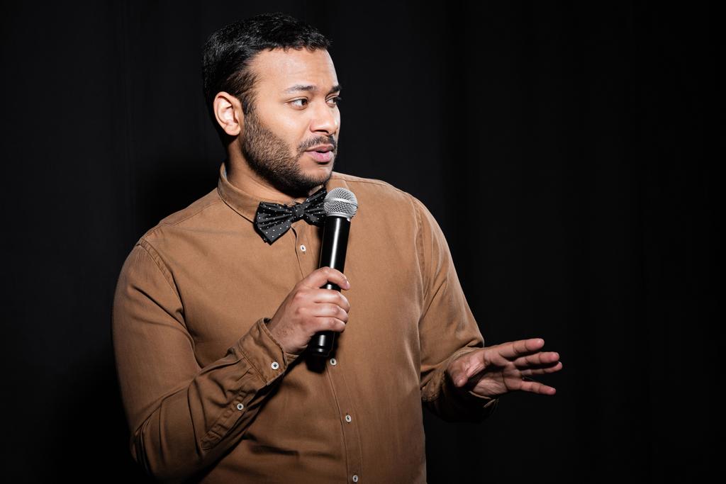 bearded indian stand up comedian in shirt and bow tie holding microphone during monologue on black  - Photo, image