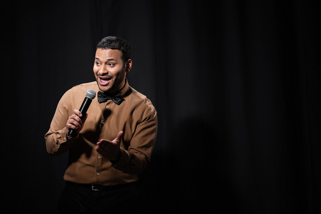 positive indian comedian in shirt and bow tie holding microphone during monologue on black  - Photo, Image