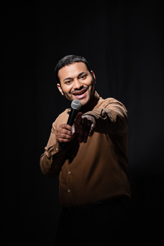 szczęśliwy indyjski komik w koszuli i muszki trzymając mikrofon i wskazując palcem podczas monologu na czarny  - Zdjęcie, obraz