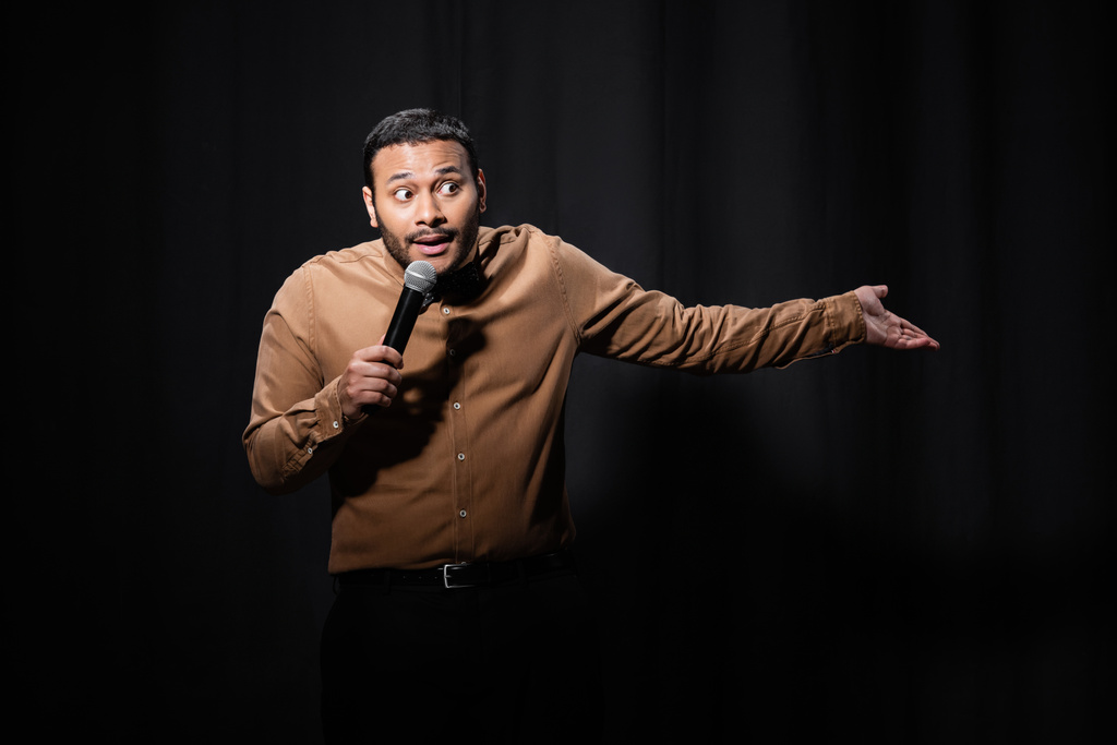 puzzled indian comedian in shirt and bow tie holding microphone and gesturing during monologue on black  - Foto, immagini