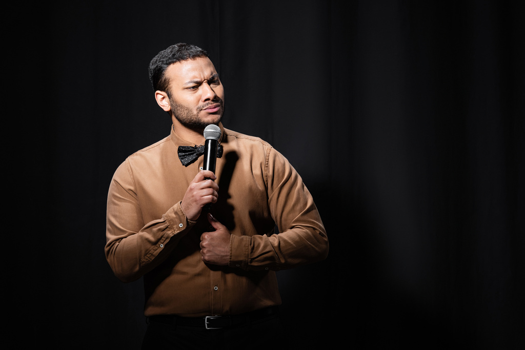 offended indian comedian in shirt and bow tie holding microphone during monologue on black  - Fotó, kép