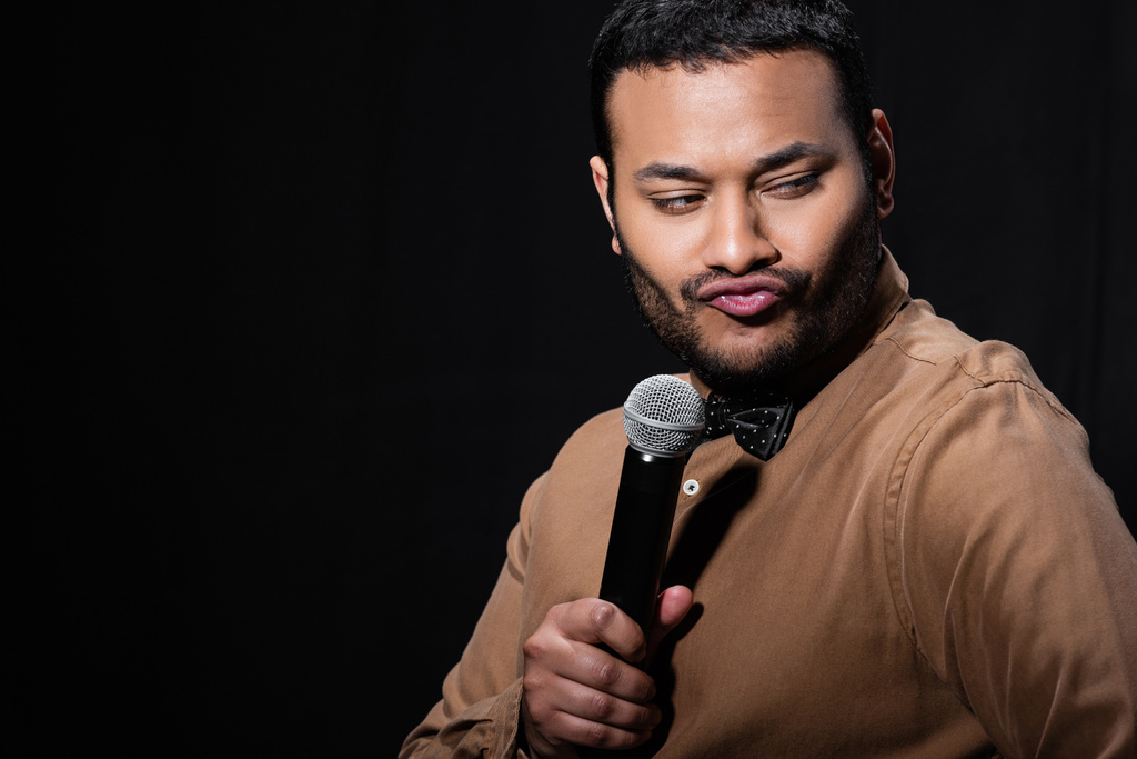 skeptical indian stand up comedian telling joke into microphone and pouting lips isolated on black  - Fotografie, Obrázek