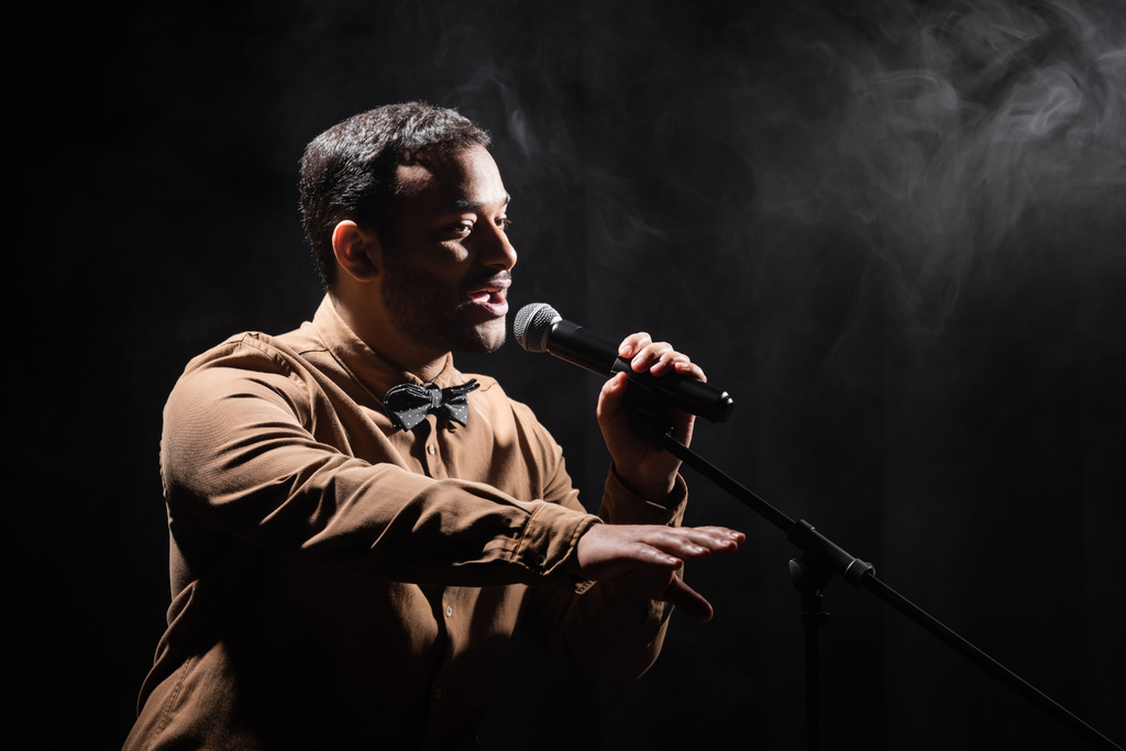 comediante indio realizando stand up comedy en el micrófono y mirando hacia otro lado en negro con humo - Foto, Imagen
