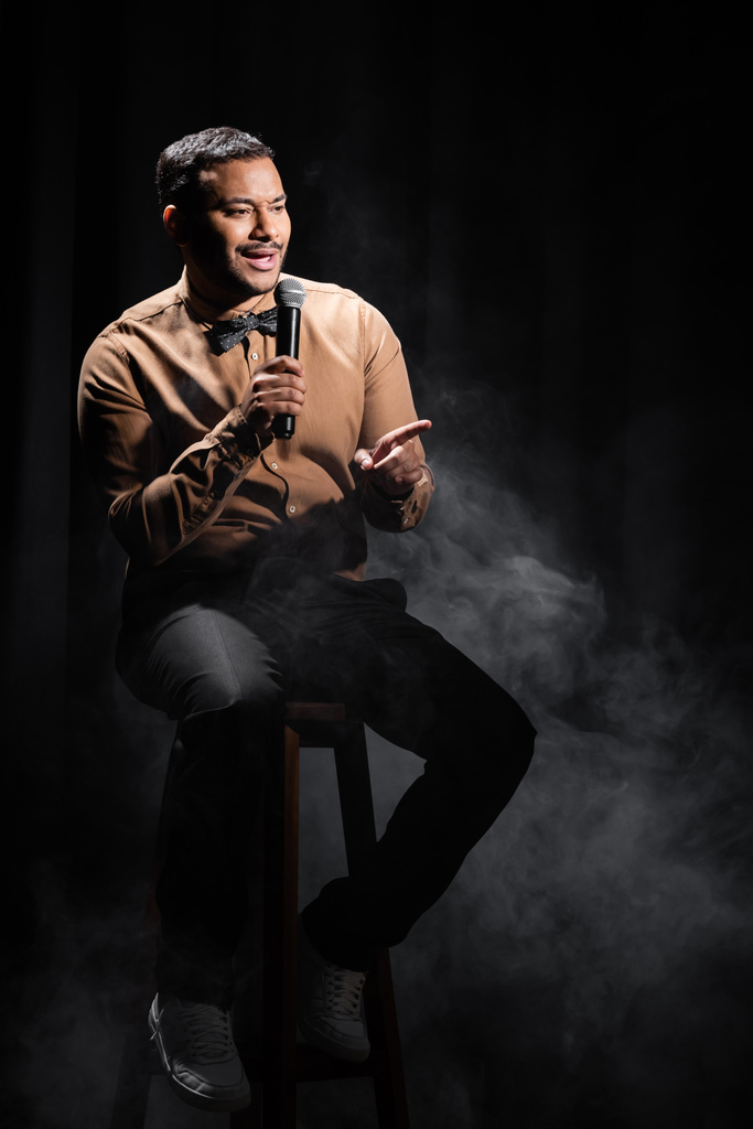 indiase komiek zitten en het uitvoeren van stand up comedy in microfoon op zwart met rook  - Foto, afbeelding