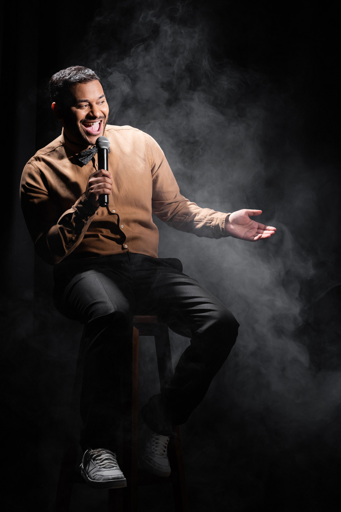 изумленный индийский комик, сидящий и исполняющий стендап-комедию в микрофон на черном  - Фото, изображение