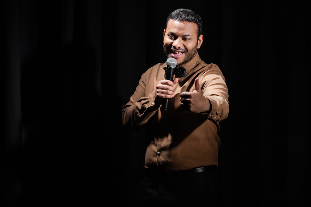 joyful middle east comedian showing like and performing stand up comedy into microphone isolated on black  - Zdjęcie, obraz