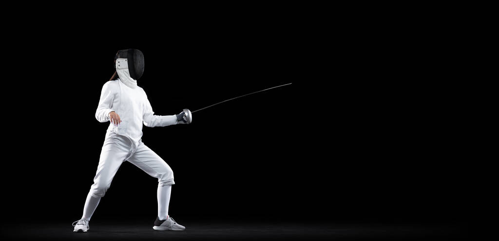 Basic stance. One female fencer in white fencing costume and mask in action, motion isolated on dark background. Sport, youth, activity, skills, achievements. Girl practicing with rapier - Foto, Imagem