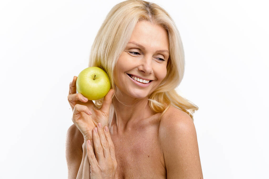 Korosodás elleni kezelés. Vidám, érett 50-es éveiben járó nő friss zöld almával és mosollyal a kezében. Középkorú modell, csupasz vállakkal pózol és félrenéz, fehér alapon elszigetelve. - Fotó, kép