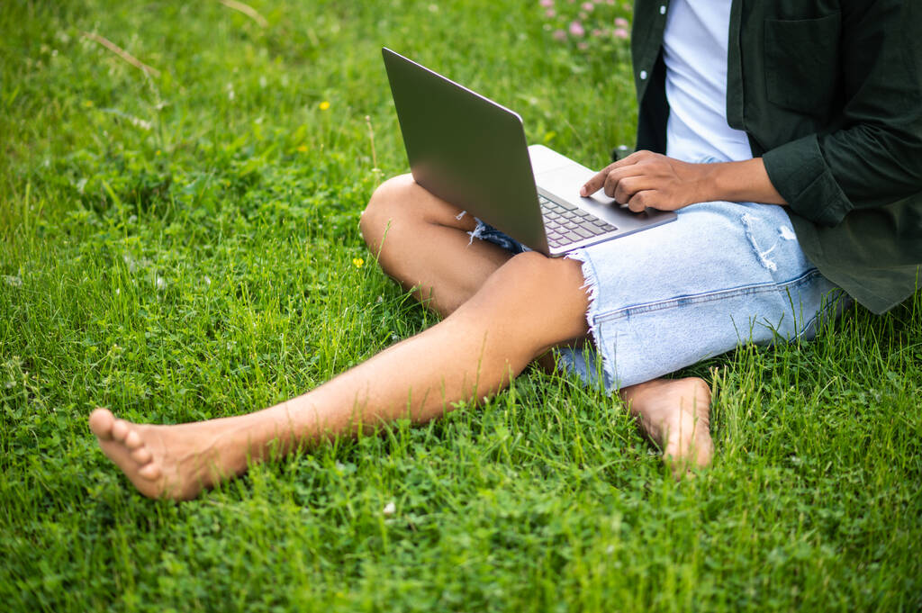 Zeit für sich selbst. Beine eines dunkelhäutigen Mannes, der am Laptop arbeitet und ohne Gesicht auf Gras in der Natur sitzt - Foto, Bild