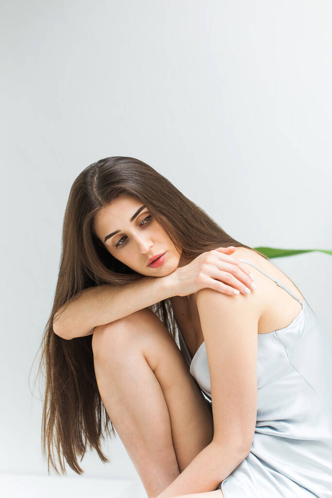 Nachdenkliche Frau mit langen dunklen Haaren, die Peignoir trägt, sitzt auf hellem Hintergrund im Studio. Elegante Dame stützt sich auf gebeugte Knie. Schönheit und Mode - Foto, Bild