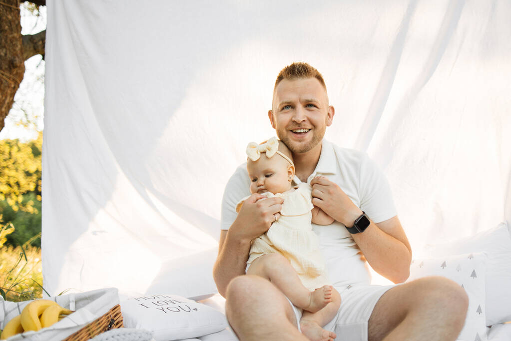 Blond haired cheerful happy man relaxing on white blanket with curtain in park with little baby girl while looking at camera. Family summer concept. - Photo, Image