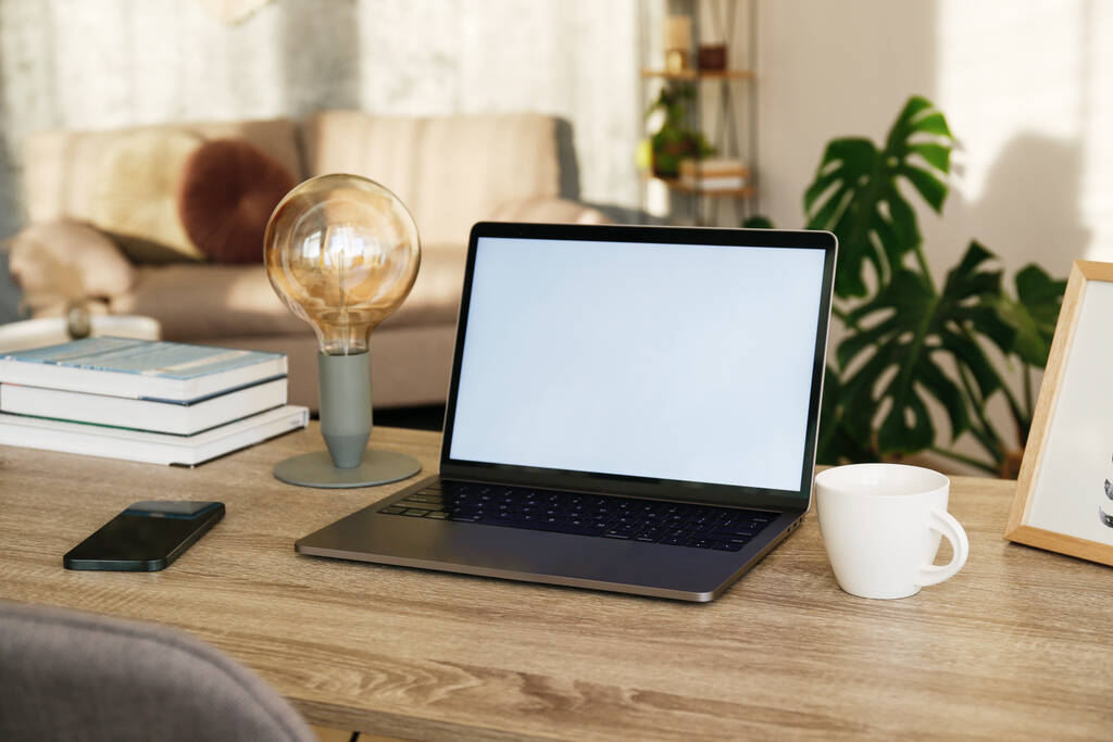 Blank screen laptop, cell phone and a cup on wooden table. Cozy lofty office with house plants full of natural sunlight. Close up, copy space, background. - Photo, Image