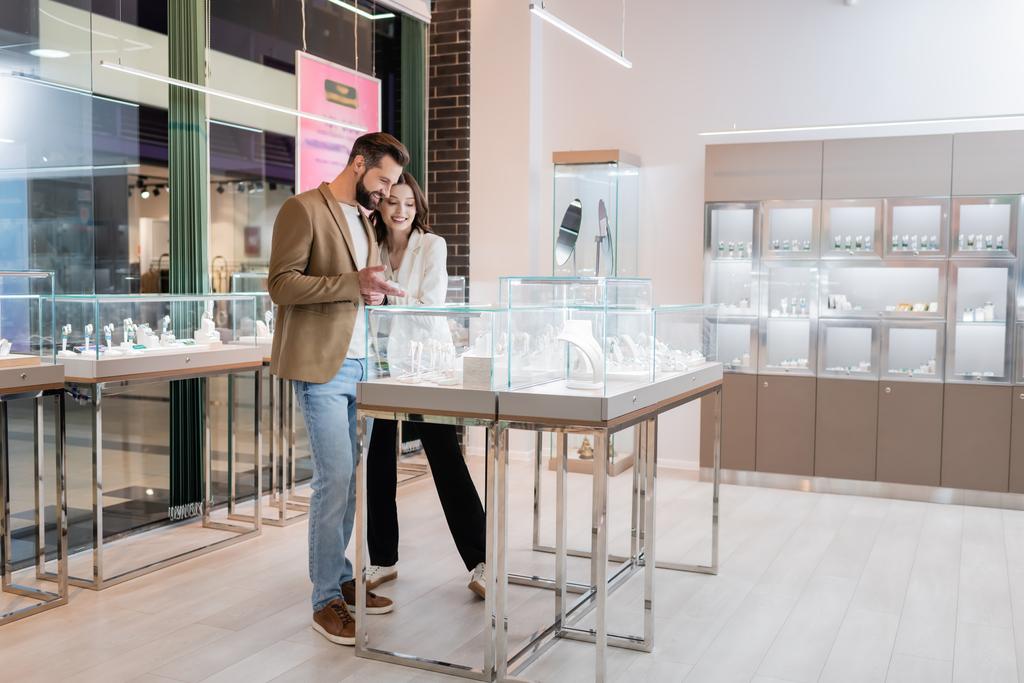 Bearded man pointing with hand at jewelry near brunette girlfriend in store  - Fotó, kép