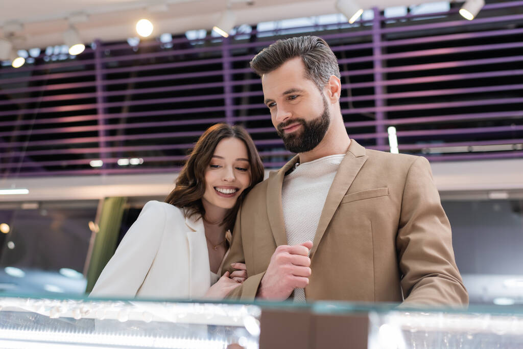 Smiling woman hugging boyfriend near blurred showcase in jewelry store  - Fotografie, Obrázek