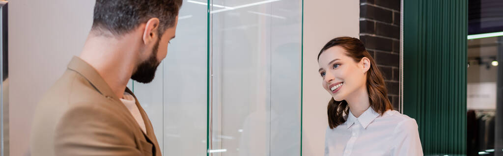 Θετικός πωλητής κοιτάζοντας θολή πελάτη κοντά βιτρίνα γυαλί σε κοσμηματοπωλείο, πανό  - Φωτογραφία, εικόνα