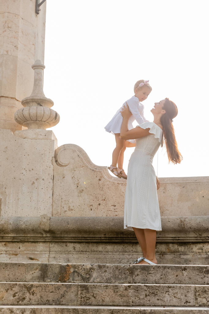 Smiling mother in dress holding daughter on stone stairs of Puente Del Mar bridge in Valencia - Fotoğraf, Görsel