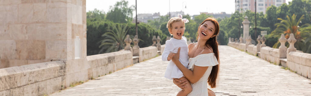 Fröhliches Baby im Sommerkleid mit Blick in die Kamera neben Mama auf der Brücke Puente del Mar in Valencia, Banner  - Foto, Bild