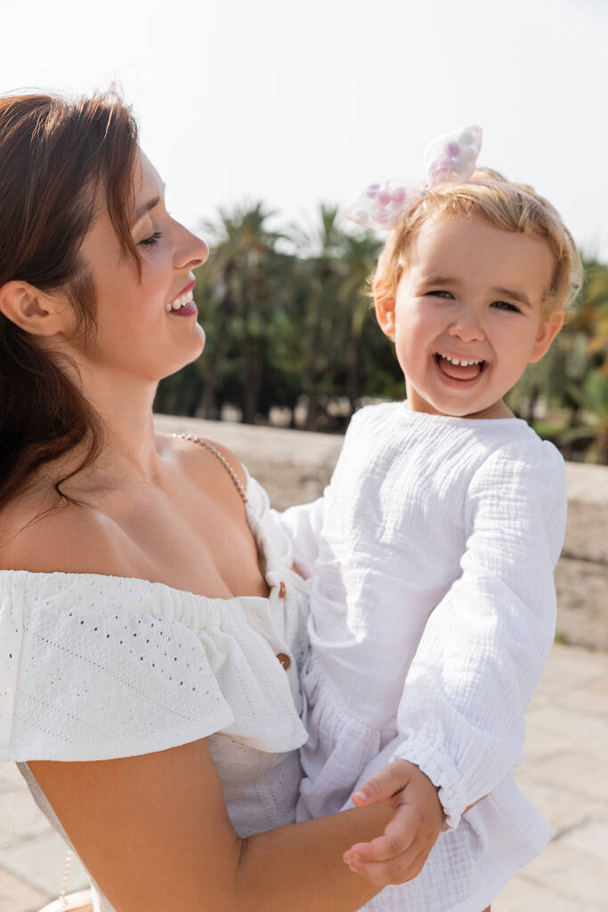 Portrait of baby girl in white dress looking at camera near mom in Valencia - Photo, Image